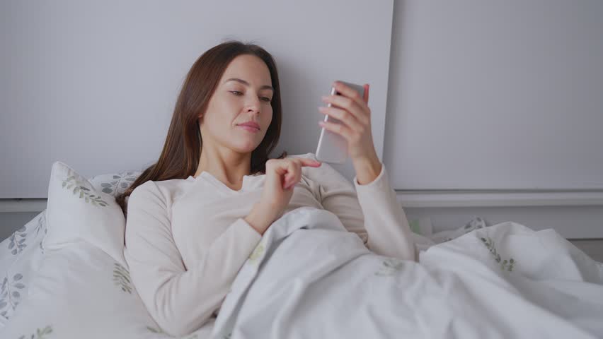 A young woman lies in bed covered with a sheet, browsing her smartphone with a focused expression, representing technology use in everyday life. Royalty-Free Stock Footage #3470456079