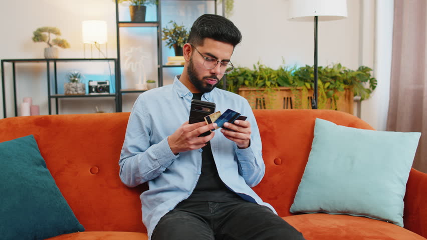 Happy young Indian man doing payments online purchase shopping in internet store. Smiling Hispanic guy holding banking credit cards involved in verification process in mobile app on sofa at home.
