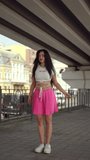 Vertical video. A girl with long black hair dances a Latin dance under a city bridge on a summer day outdoors. The girl looks at the camera and smiles while dancing