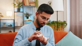 Indian young bearded man taking medicine pill with water sitting on cozy sofa at home. Hispanic guy takes medication prescribed by his physician. Medicine, health care, pharmacy and people concept. - Powered by Shutterstock - Get 15% off with code: PIKWIZARD15