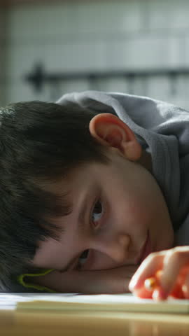 Vertical video. Tired little schoolboy kid lying on table desk writing homework, boring class lesson drawing in notebook at home.