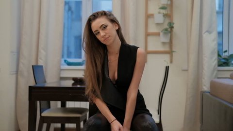 Sensual young woman with long brown hair in a black suit posing at camera in front of the apartment.