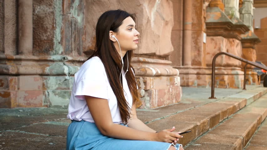 Toned footage of smiling young girl sitting on stone staircase and listening music on mobile phone