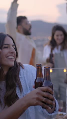 Vertical. Group of young diverse people dancing excited and toasting with beers at rooftop barbecue party. Laughing friends jumping and having fun drinking at event in sunset summer. Slow motion
