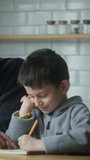 Vertical video. Young father help little schoolboy pupil with homework at home. Dad helping to son with writing while sitting in the kitchen. Adult parent or tutor with kid boy learning. Private educa