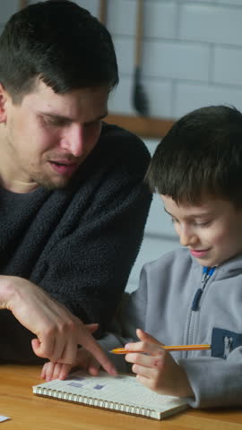 Vertical video. Young father help schoolboy with homework at home. Dad helping to son with writing while sitting in the kitchen. Adult parent or tutor with kid boy learning.