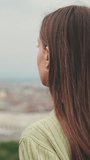 Vertical video, Young woman with brown hair wearing an olive green sweater looks at the city from the observation deck