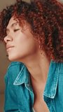 Vertical video, Close-up of contemplative young woman with curly hair wearing denim shirt looking through window while relaxing on sofa in cozy living room