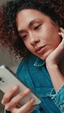 Vertical video, Close-up of young smiling woman with curly hair wearing denim shirt looking at mobile phone while sitting on sofa in cozy living room