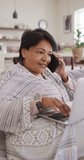 Vertical video of smiling senior biracial woman talking on smartphone in living room. healthy and active retirement at home.