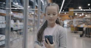 Happy child making online video call discussing food in supermarket using smartphone talking. Modern technology and shopping concept.