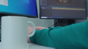 A person is working at a computer with a white cup of coffee on a desk in the foreground. A man's hand takes it and places it back.. Concept of productivity and focus, as the person is working on thei