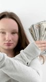 rich teenager girl waving a fan of 100 euros and 100 dollar bills standing on white background. smiling in a sports gray sweater loose brown hair surprise calm rich child
