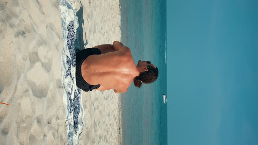 Vertical video man sitting under sun on beach with golden sand, enjoying view ocean. Traveler enjoying moment on beach, breathing in fresh sea air. Beach gives feeling of peace and unity with nature.