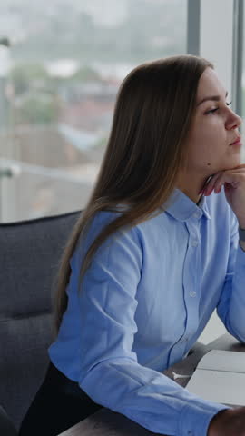 Young female employee sitting at desk in the office thinking over some issues. Long-haired starts to talk to camera smiling and waving hands. Vertical video Royalty-Free Stock Footage #3470915895