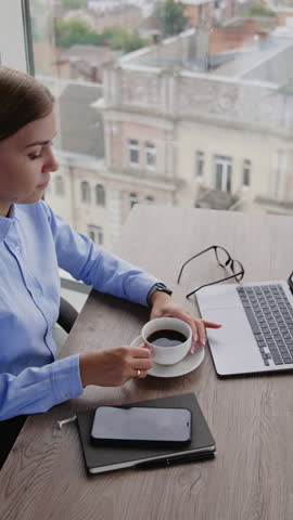 Blonde female sitting at desk in office holding a cup of coffee. Calm relaxed lady enjoying lunch time at work place. High angle view. Vertical video Royalty-Free Stock Footage #3470916861