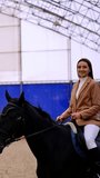 Happy smiling lady taking lessons of horse riding. Beautiful long-haired brunette rides on a black horse slowly. Manege backdrop. Vertical video