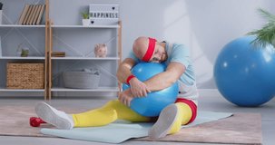 Funny young man wearing bright sportswear sitting on yoga mat and sleeping on fit ball tired and resting from sport exercises at home. Fitness, health and home workout concept. 4k video.