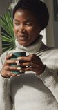 Vertical video of african american woman drinking coffee and looking outside window. leisure, relax and spending time at home.