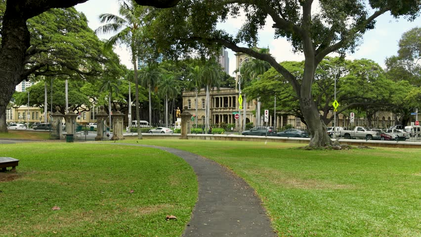 footage along a footpath at the Iolani Palace with lush green trees and grass, buildings in the skyline, the King Kamehameha Statue and cars driving on the street in Honolulu Hawaii USA