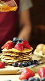 Man in apron and black gloves pouring honey over pancakes with cream, strawberries and blueberries on wooden board, warm light, vertical video