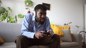 An African American man grins broadly while gaming at home, using a wireless controller and headset, fully immersed in the interactive experience. Enthusiastic Gamer Engaged in Play with Wireless Head