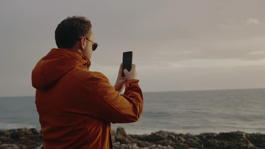 Back view on man hold smartphone and take picture of beautiful seascape. Young male walking on ocean coast and shooting video of nature using cellphone camera