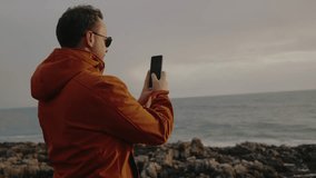 Back view on man hold smartphone and take picture of beautiful seascape. Young male walking on ocean coast and shooting video of nature using cellphone camera - Powered by Shutterstock - Get 15% off with code: PIKWIZARD15