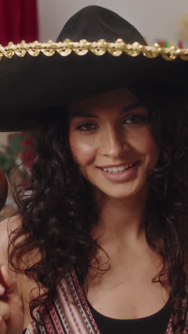 Medium vertical zoom out portrait shot of beautiful Mexican girl in sombrero dancing with maracas, smiling, while having fun at Cinco de Mayo house party