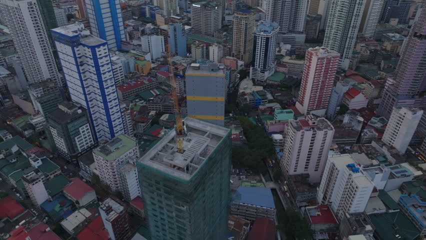 High angle view of construction of tall building with crane on top. Tilt up reveal residential towers and sunset cityscape. Manila, Philippines