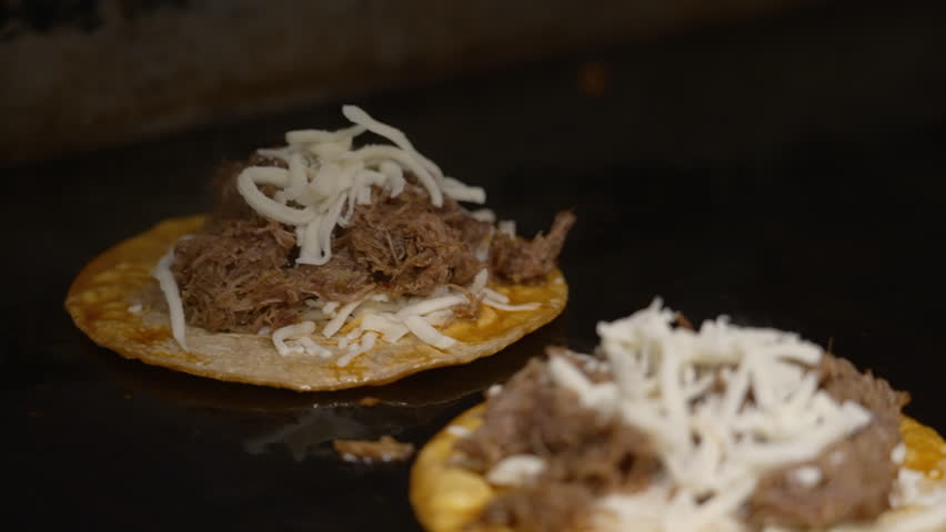 Shredded cheese added to birria tacos cooking on flat top grill, black gloved hand sprinkles grated quesadilla cheese to sizzling tacos, slow motion slider close up 4K