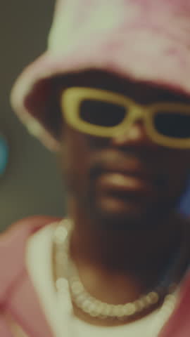 Vertical zoom-in closeup portrait of stylish African American hipster guy posing for camera indoors wearing pink plushy bucket hat and yellow glasses