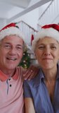 Vertical shot of a senior Caucasian couple having a video call, wearing Santa Clause hats. Seniors, technology, and Christmas concept.