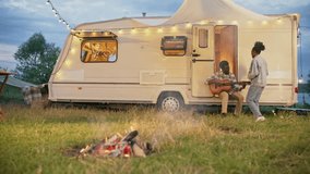 African American couple enjoying romantic weekend by white caravan with glowing garland. Attractive man playing on guitar to his girlfriend. Woman holding drink and dancing at music. Flame in nature. - Powered by Shutterstock - Get 15% off with code: PIKWIZARD15