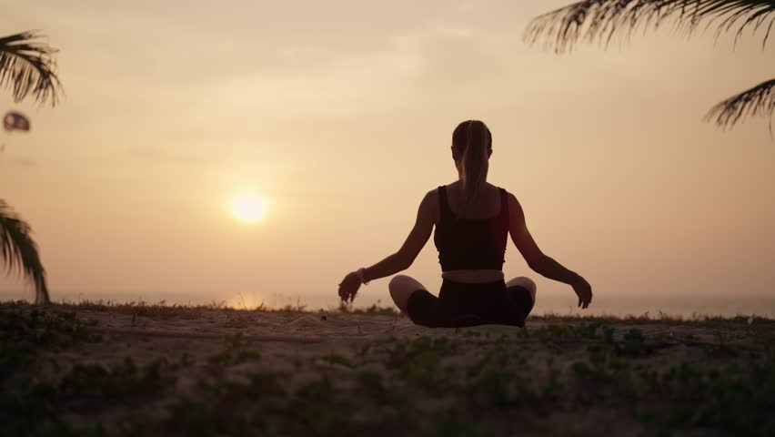 Woman practices yoga on ocean at sunset. Silhouette sporty yoga girl doing yoga pose asana exercise stretching at sunset on beach in nature close up. Peace, morning routine, relax on sea concept.
