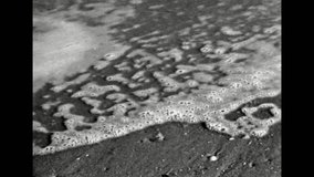 Close-up, monochrome film of sea foam on sand. Seascape sky horizon over sea. Abstract tide of sea waves on sandy coast. Ocean beach landscape. Overscan footage. Vintage black white retro archive 1980