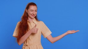 Redhead woman showing thumbs up and pointing right empty place, advertising area for commercial text, copy space for goods promotion advertisement. Girl isolated on blue studio background. Lifestyles - Powered by Shutterstock - Get 15% off with code: PIKWIZARD15
