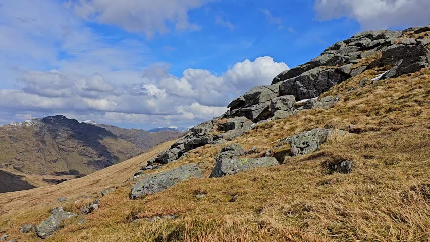 High atop the mountain, a captivating video reveals a vast valley, distant peaks, rocky formations, and shifting clouds against green-yellow grassy expanse.