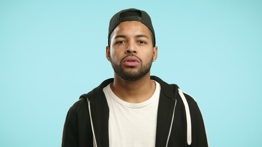 A pensive young man with braces on his teeth and wearing a baseball cap gestures for silence, placing his finger over his lips on a soft blue background. Camera 8K RAW. 