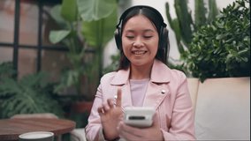 An Asian young woman in a cafe selects songs on her smartphone, enjoying music with her noise-canceling headphones amidst plants.