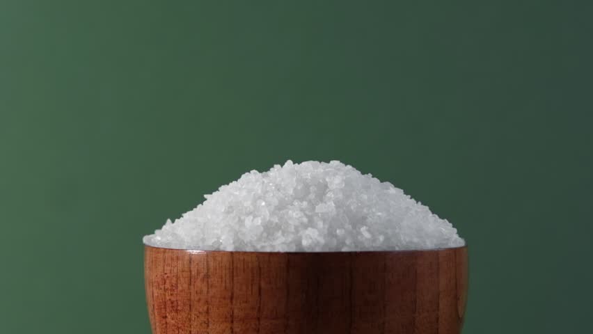 White table salt granules falling into a pile. Wood bowl. Gray color studio background. Condiment particles dropping. Clean cosmetic bath salt pouring footage. Closeup of grained sea salt pieces hill