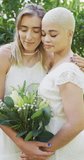 Vertical video of happy diverse female couple with bunch of flowers embracing in garden. wedding day and celebration concept.