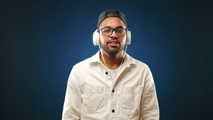 A cheerful biracial man with braces enjoys his favorite tunes, waving American flags with pride and excitement against a dark blue background. Camera 8K RAW. 