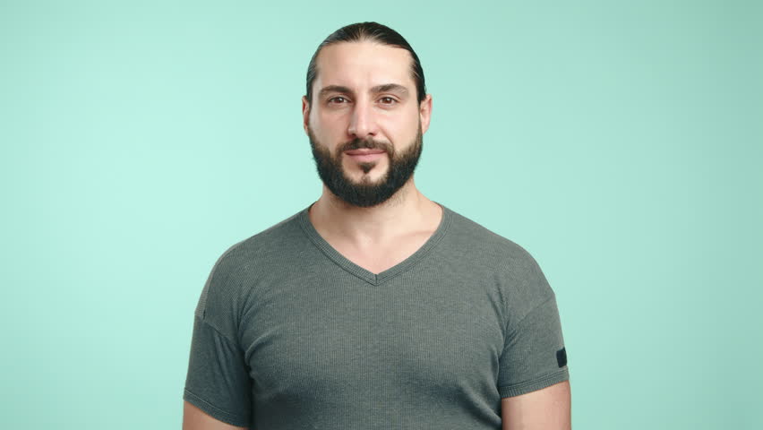 A friendly businessman with long hair and a casual grey t-shirt greets warmly, waving his hand with a broad smile, evoking a sense of welcoming and approachability on a teal background. Camera 8K RAW.