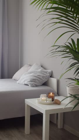 Home decor, stylish interior. Burning candles on marble tray standing on white badside table in bedroom.  Bed with grey blanket and pillows, howea palm plant. Vertical video.