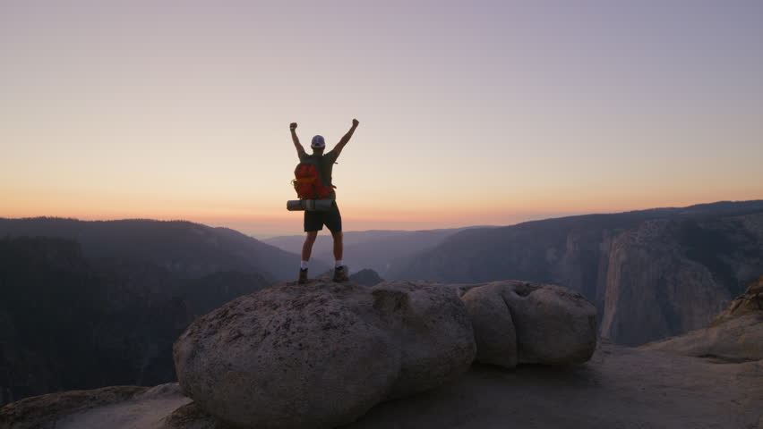 Amazing view of dark mountain range with orange skyline, happy male hiker on a stone on a summit raising hands up with delight, overwhelmed with emotions. High quality 4k footage