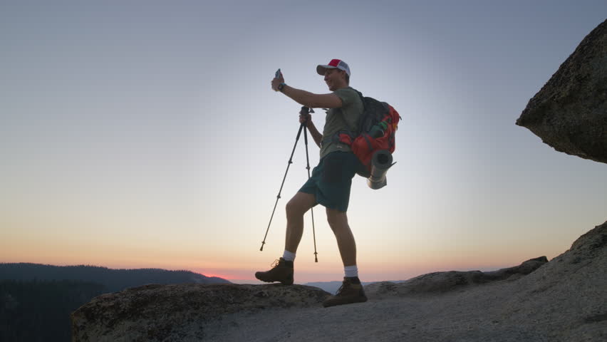 Young male backpacker with hiking gear having video conference on smartphone standing on the peak of mountain showing sweeping panoramas,of Yosemite park. High quality 4k footage