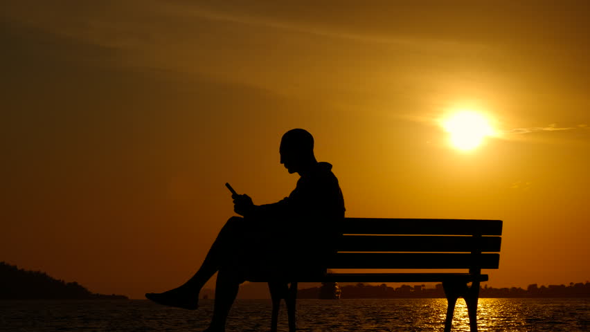 Relaxing man with phone at nightfall. A man relax with his phone on bench by the lake during nightfall time.