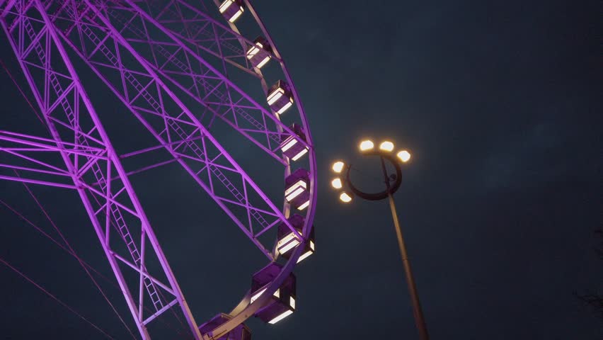 Purple ferris wheel medium shot, ground level side shot in night time, street lamp. Lyon ferris wheel, place Bellecour, France. Cloudy night sky.