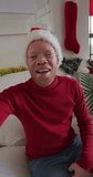 Vertical shot of an Albino man having a video call, wearing a Santa Clause hat, smiling at the camera. Christmas concept.